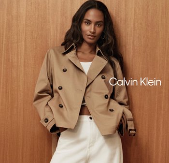 Mujer de pelo largo y oscuro que lleva una chaqueta beige de doble abotonadura sobre un top corto blanco y pantalones blancos, posando frente a un fondo de madera.