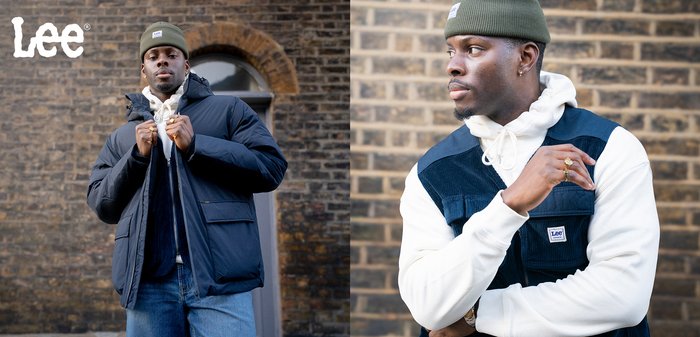 Mężczyzna w zielonej czapce, białej bluzie z kapturem, niebieskiej kamizelce i ciemnoniebieskiej kurtce pozuje na tle ceglanej ściany z logo Lee w rogu.