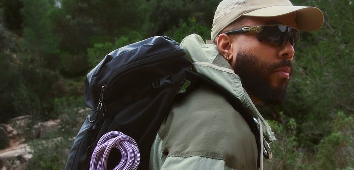 Homme portant des lunettes de soleil et une casquette porte un sac à dos noir avec une corde violette enroulée dans un cadre forestier.
