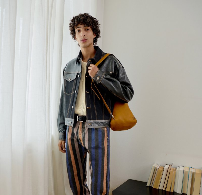 Jeune personne aux cheveux bouclés porte une veste en cuir noire, un pantalon rayé et tient un sac à bandoulière marron sur un fond uni.