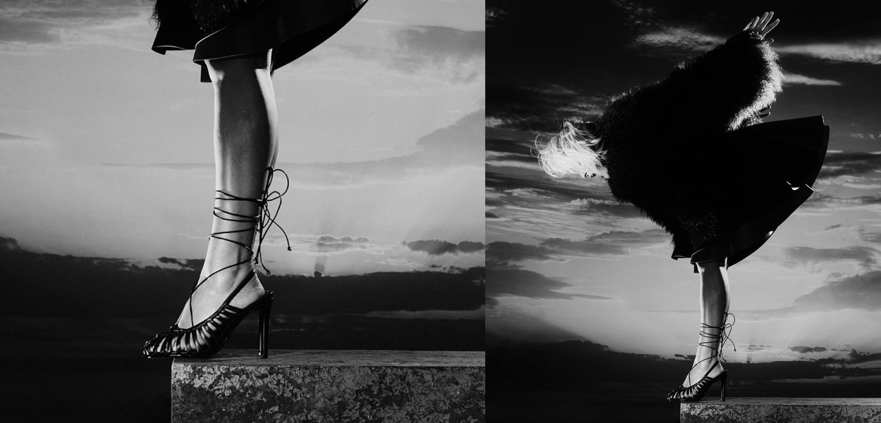 Czarny but na wysokim obcasie z otwartym noskiem i krzyżującymi się paskami. Modelka nosi czarną spódnicę oraz fakturowaną czarną odzież wierzchnią.