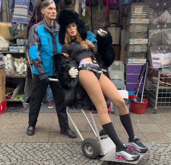 En man i en blå jacka skjuter en vagn med en lutande figur som bär en svart pälsrock och sportiga leggings, vilket visar upp kontrasterande texturer.