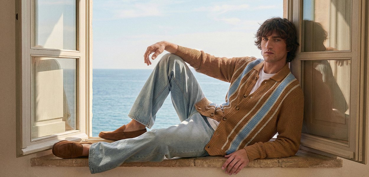 Joven con ropa casual recostado en el alféizar de piedra de una ventana con contraventanas abiertas, con vista al mar tranquilo y cielo despejado.