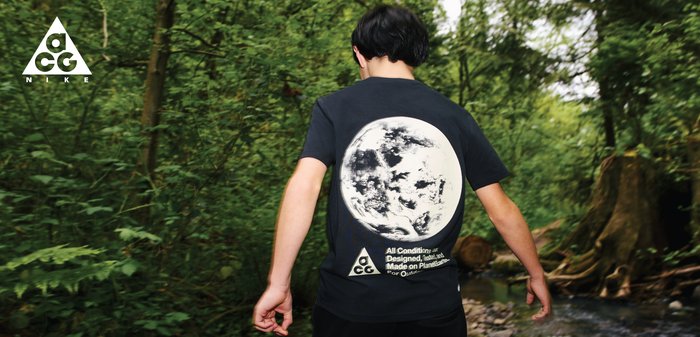 Personne portant un t-shirt noir Nike ACG avec un graphisme de lune marchant à travers une forêt verte près d'un ruisseau.