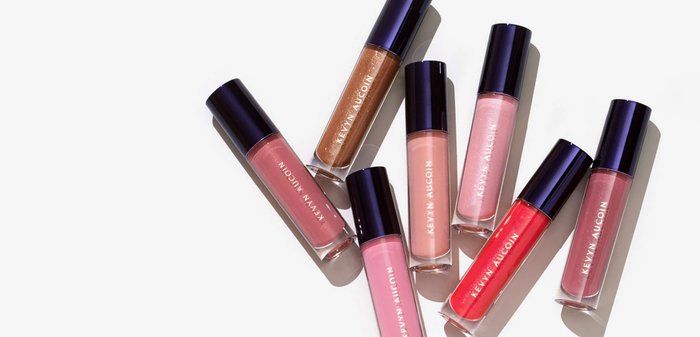 Sette tubetti di lucidalabbra disposti a angoli diversi. I colori includono rosa, corallo e un bronzo metallico. Basi trasparenti con tappi blu scuro.