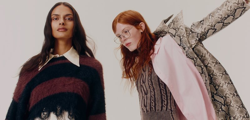 Δύο μοντέλα φορούν διαφορετικές εμφανίσεις: ένα καρό πλεκτό σε μπορντό και μαύρο με κρόσσια και ένα παλτό με σχέδιο φιδιού πάνω από μια υφασμάτινη μπλούζα με ροζ πουκάμισο.