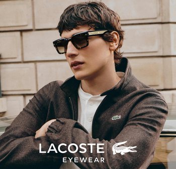 Młoda osoba w okularach przeciwsłonecznych w kolorze żółwia i ciemnej kurtce z logo Lacoste, stojąca na zewnątrz w tle kamiennej ściany.