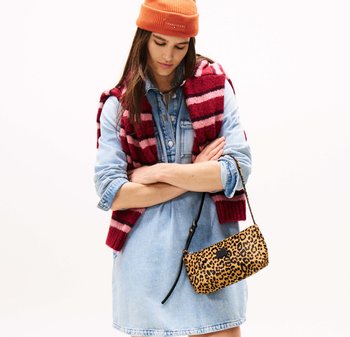 Torebka typu crossbody w leopardzie wzorze z czarnym paskiem, noszona na tle niebieskiej sukienki dżinsowej i założona na prążkowany bordowy i różowy kamizelkę. Na głowie pomarańczowa czapka.