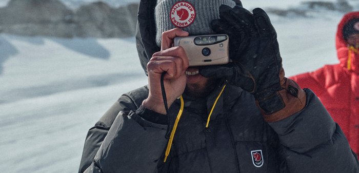 Beige Filmkamera, die von einer Person gehalten wird, die eine graue Daunenjacke mit rotem Logo und schwarzen Handschuhen trägt, mit Schnee im Hintergrund.