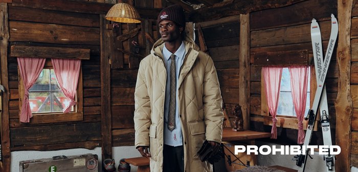 Beige quilted puffer jacket, long sleeves, side pockets, paired with a button-up shirt and tie, worn with brown gloves in a rustic wooden setting.