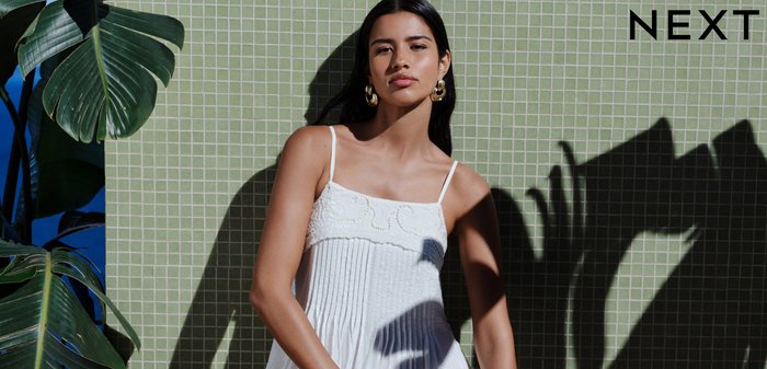 Mujer con vestido blanco sin mangas que sostiene un bolso tejido, de pie contra una pared verde con azulejos y grandes sombras de plantas detrás de ella.