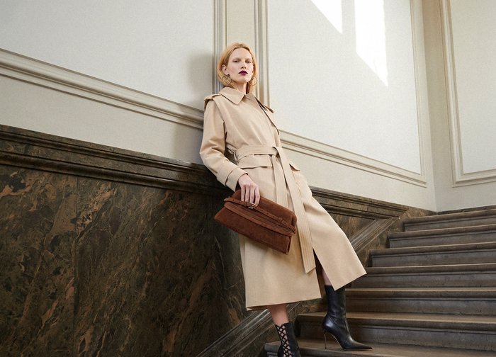 Femme en trench-coat beige et bottes à talons noires tenant une pochette marron en s'appuyant sur une rampe d'escalier en marbre dans un intérieur lumineux.