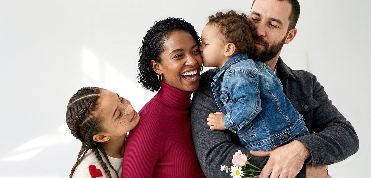 Kind in Jeansjacke küsst lächelnde Frau im roten Rollkragenpullover, Mann hält Kind und Blumen, Mädchen mit geflochtenem Haar lehnt sich an die Schulter der Frau.