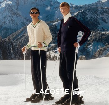 Dos esquiadores están de pie en la nieve: uno lleva un suéter de punto tipo cable color crema con detalles verdes y el otro un suéter azul marino con una franja roja.