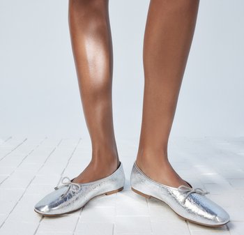 Zilveren platte schoenen van metallic materiaal met een ronde neus, een delicate strikdetail en een platte zool, geplaatst op lichte tegels.
