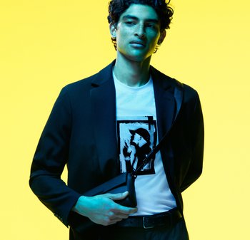 Czarna marynarka na białym t-shircie z czarnym nadrukiem. Modelka trzyma małą czarną torbę, na tle żywej żółtej ściany.