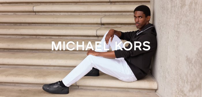 Mężczyzna w czarnej kurtce, białych spodniach i czarnych butach siedzi na beżowych kamiennych schodach przy ścianie z tekstem "MICHAEL KORS".