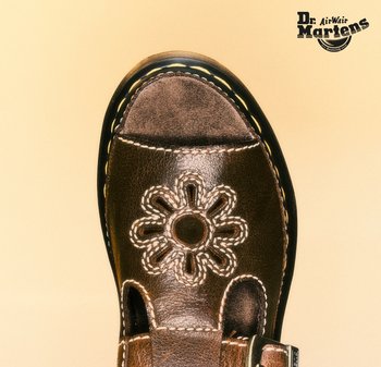 Bruine leren sandaal met bloemvormige uitsnede en gele stiksels, gebrandmerkt met het Dr. Martens-logo op een beige achtergrond.