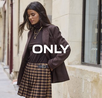 Vrouw in een bruine blazer, een donkere coltrui en een geruite rok houdt een koffiekop vast terwijl ze langs een stadsstraat wandelt naast een stenen gebouw.