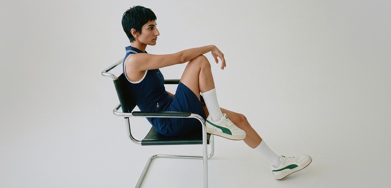 Lyhyttukkainen henkilö, joka pukeutunut hihattomaan mekkoon ja tennareihin, istuu sivuttain modernissa metallikehyksisessä tuolissa yksinkertaista taustaa vasten.