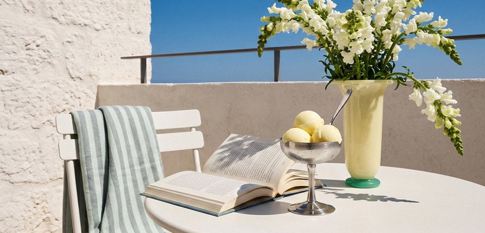 Λευκό στρογγυλό τραπέζι με ανοιχτό βιβλίο, μπολ με σορμπέ λεμονιού και κουτάλι, κίτρινο βάζο με λευκά λουλούδια και καρέκλα καλυμμένη με ριγέ πετσέτα σε εξωτερικό χώρο.