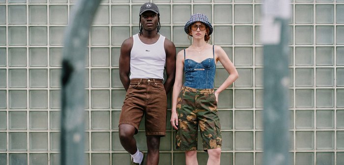 Deux jeunes adultes se tiennent devant un mur en verre texturé, vêtus de vêtements d'été décontractés incluant des shorts, un débardeur et des chapeaux.