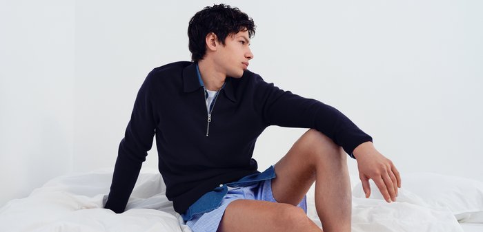 Junger Mann sitzt auf dem Bett, trägt ein dunkles Langarmshirt und blaue Shorts, schaut nach rechts vor einem schlichten weißen Hintergrund.