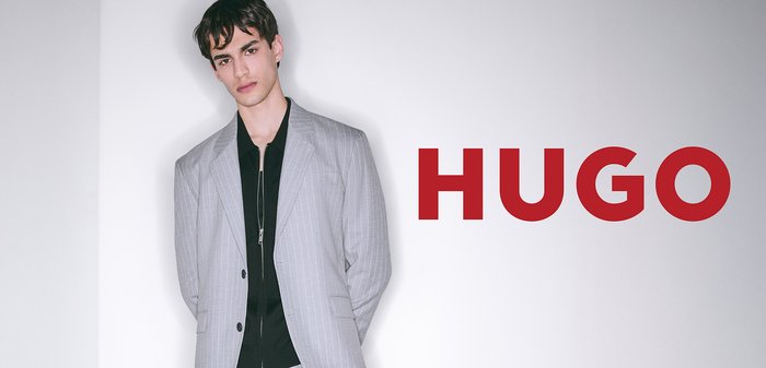 Hellgrau gestreifter Blazer über einem schwarzen Hemd, mit Reißverschluss und taillierter Passform. Auffälliger roter "HUGO"-Schriftzug rechts.