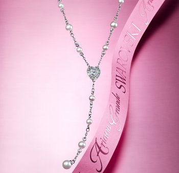 Silberne Halskette mit einem herzförmigen Kristall-Mittelstück, akzentuiert mit runden Perlen, vor einem rosa Hintergrund mit einem rosa Band.
