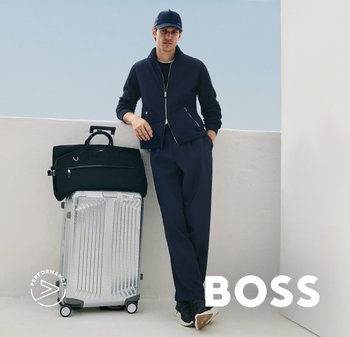 Man in marineblauwe casual kleding en pet leunt op zilverkleurige harde koffer met zwarte reistas tegen een witte muur, "BOSS"-logo zichtbaar.