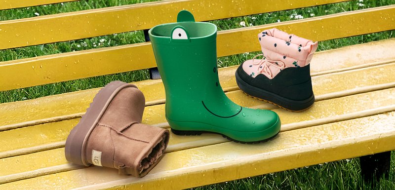 Trois paires de bottes sur un banc jaune : une botte en caoutchouc verte de pluie avec un sourire, une botte en suède marron, et une botte isolée rose et noire avec des accents floraux.