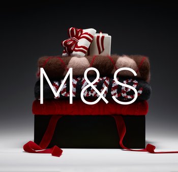 Stos swetrów w różnych odcieniach czerwieni, brązu i czerni z wzorami. Na górze, na pudełku prezentowym, znajduje się biała świeca ozdobiona czerwoną wstążką.