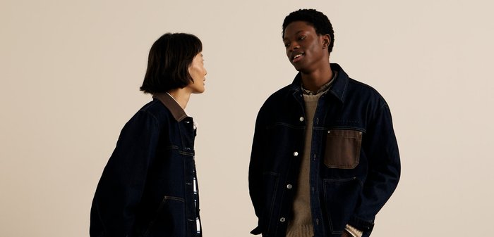 Dos personas con chaquetas de mezclilla oscura, una con un detalle de bolsillo marrón, contra un fondo beige. La tela muestra una textura clásica y resistente.