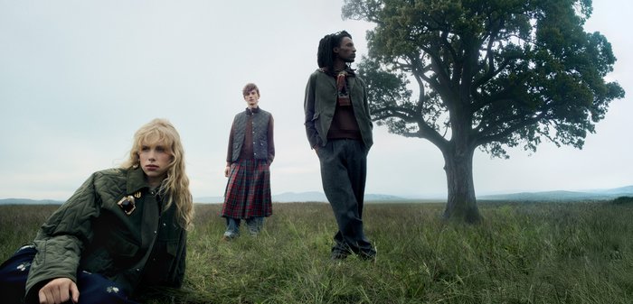 Trois mannequins posent dans un champ herbeux. L'une porte une veste matelassée vert olive avec un accent doré. Une autre porte un gilet marron associé à une jupe à carreaux. Le troisième mannequin porte une tenue grise avec une écharpe, debout à côté d'un grand arbre.