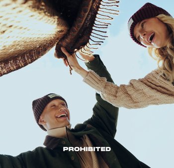 Two individuals holding a woven blanket with fringes. The background is clear blue sky. Both wear knit beanies and textured sweaters.