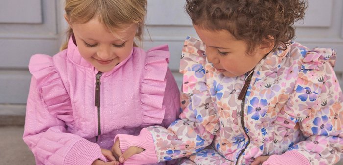 Junge trägt eine beige Jacke und ein graues Sweatshirt mit Palme- und Wellenmuster, steht im Freien neben einem Metallgeländer und einer Tür.