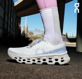 Nahaufnahme einer Person, die weiße On Running Schuhe mit CloudTec-Sohlen und weiße Socken trägt, stehend auf einer Asphaltfläche unter einer Brücke.