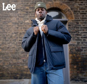 Man iklädd grön mössa, marinblå vadderad jacka, huva och jeans står mot en tegelvägg med ett valvformat fönster i bakgrunden.