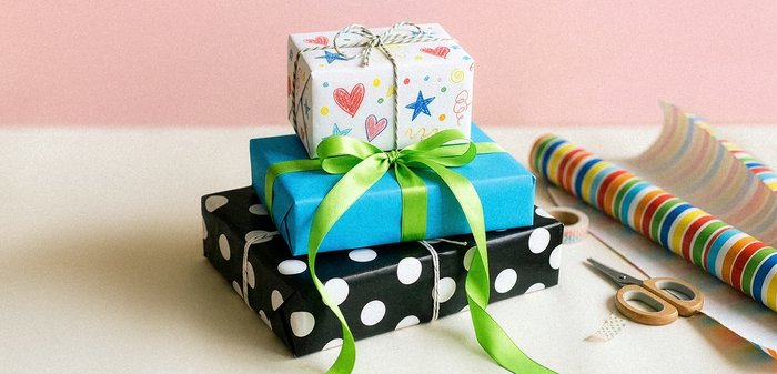 Τρία συσκευασμένα δώρα στοιβαγμένα, με πολύχρωμα σχέδια και κορδέλες, δίπλα σε ψαλίδια και ρολά περιτυλίγματος πάνω σε μια μπεζ επιφάνεια.