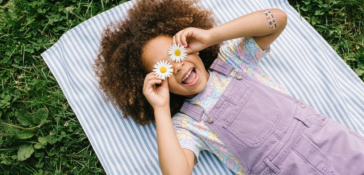 Kind liegt draußen auf einer gestreiften Decke, hält Gänseblümchen vor die Augen, lächelt mit lockigem Haar und trägt lila Latzhose und ein Blumenhemd.