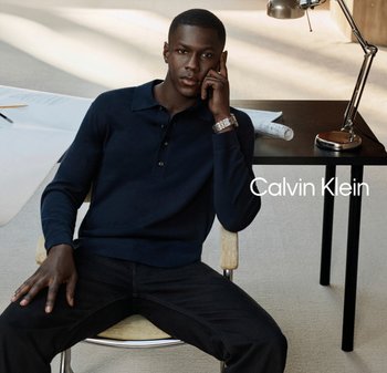 Hombre con polo azul marino de manga larga y pantalones negros sentado en una silla, apoyando la cabeza en la mano junto a un escritorio negro con lámpara y rollo de papel.