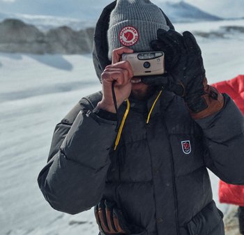 Graue Pufferjacke mit Logo, gelbem Kordelzug und lederakzentuierten Handschuhen. Person hält eine Kamera und trägt eine graue Mütze. Schneeiger Hintergrund.