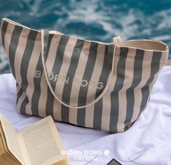 Randig grå och beige Björn Borg-tygväska på en vit handduk bredvid en öppen bok och en telefon med hörlurar vid vattnet.