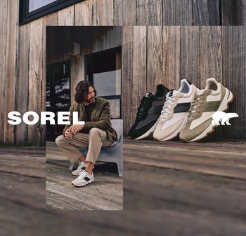 Ein Mann trägt beige Hosen und weiße Turnschuhe und sitzt auf einer Bank vor einem Holzgebäude; in der Nähe sind drei Paar Turnschuhe ausgestellt.