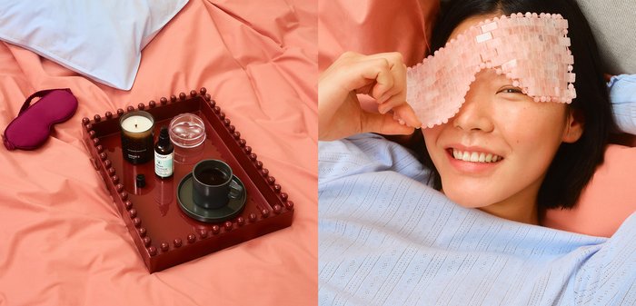 Masque de sommeil texturé rose poudré, plateau bordeaux avec des accents arrondis, une bougie, des huiles essentielles et une tasse noire sur une literie corail clair.