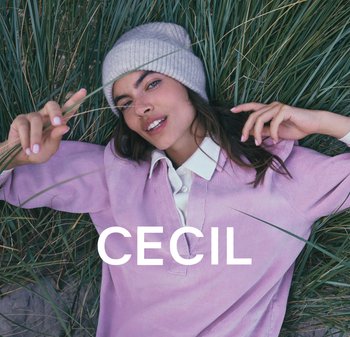 Rosa Cord-Hemd mit einem weißen Kragen, getragen unter einer hellgrauen Strickmütze. Model liegt auf grünem Gras, Hand erhoben zwischen Grashalmen.