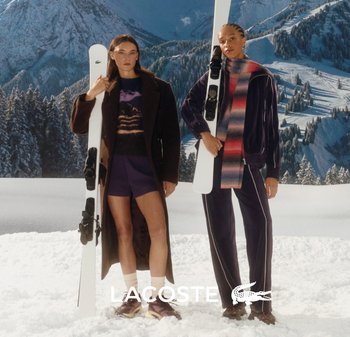 Dos modelos están de pie en la nieve, sosteniendo esquís. Uno lleva un abrigo marrón sobre un suéter estampado y pantalones cortos; el otro viste un chándal morado con una bufanda multicolor.
