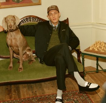 Une personne portant une veste de varsity noire et un pull gris est assise sur un canapé vert à côté d'un chien marron clair, avec un échiquier à proximité.