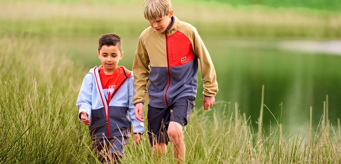 Zwei Jungen gehen durch hohes Gras in der Nähe eines Gewässers, beide tragen Jacken und konzentrieren sich auf ihren Weg.