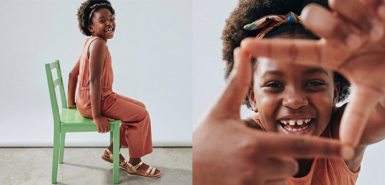 Una niña joven con ropa naranja se sienta en una silla verde sonriendo, haciendo una forma de marco con sus manos en primer plano.